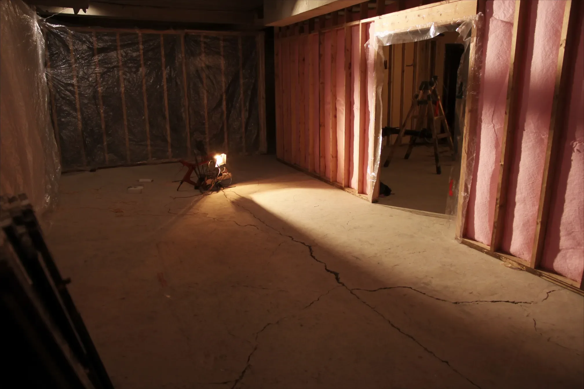 A home wine cellar under construction: framed stud walls packed with insulation, clear plastic vapor barrier being stapled over the studs, and a partially installed wood door frame in the opening.
