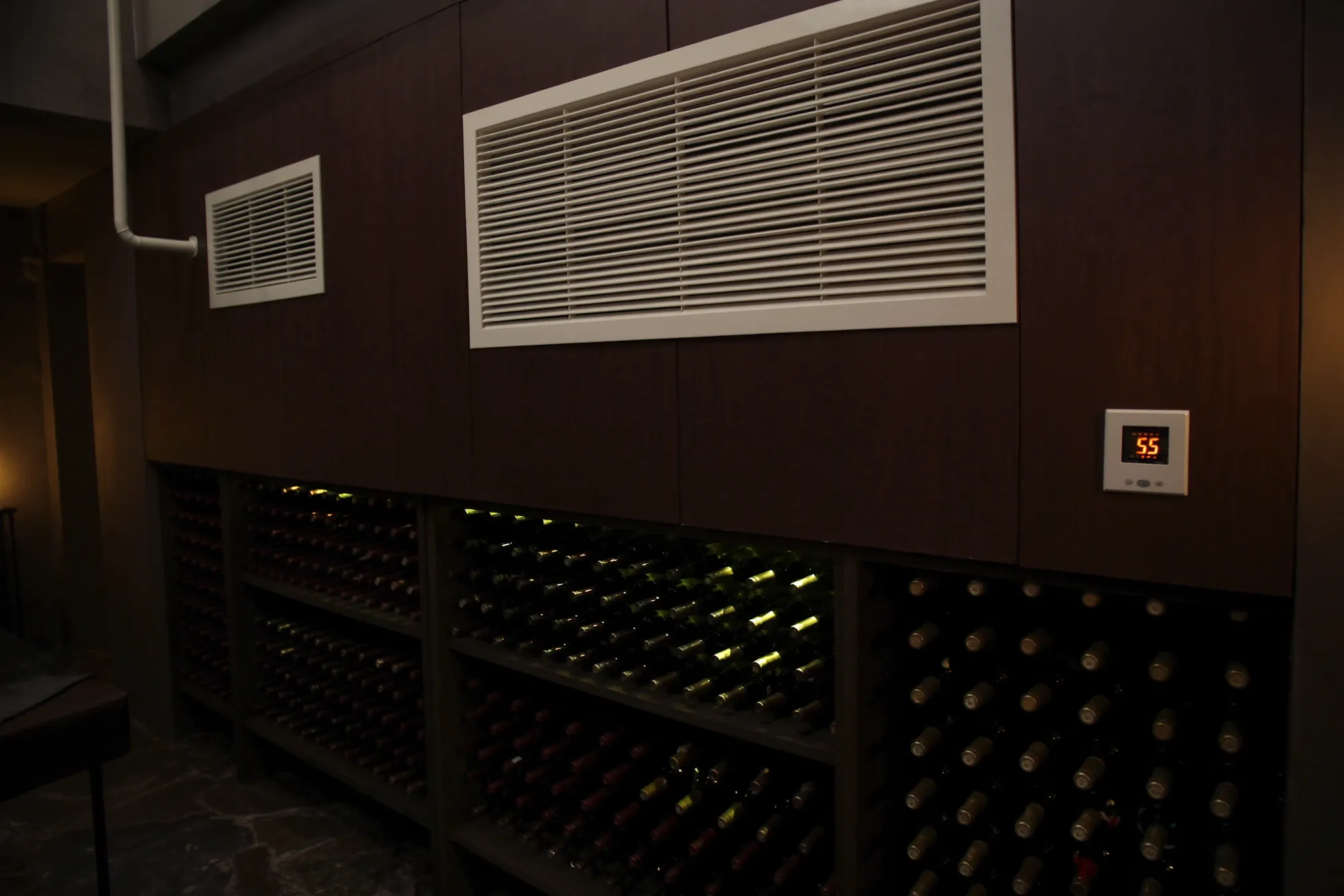 A wine cellar with ducted cooling: white supply and return grilles set high in a dark paneled wall above wooden racks, and a wall thermostat reading 55.