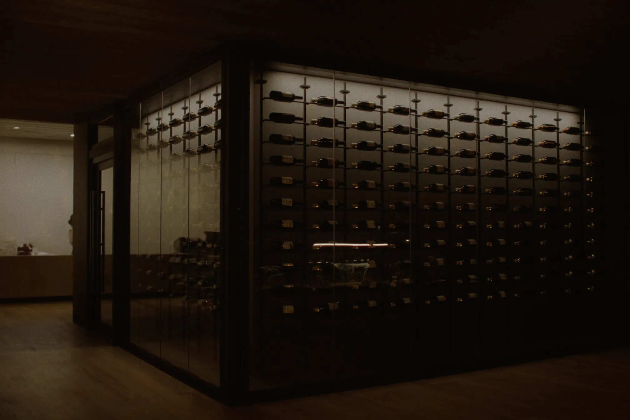 A glass-enclosed wine cellar at dusk, with full-height insulated glass walls and backlit metal racks holding bottles label-out.