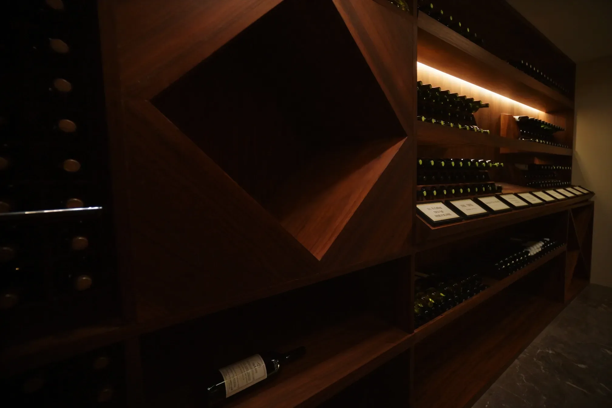 A custom home wine cellar wall of dark wood racking that combines individual bottle slots, a diamond bin, and a label-forward display row, lit by warm LED strips along the shelves.