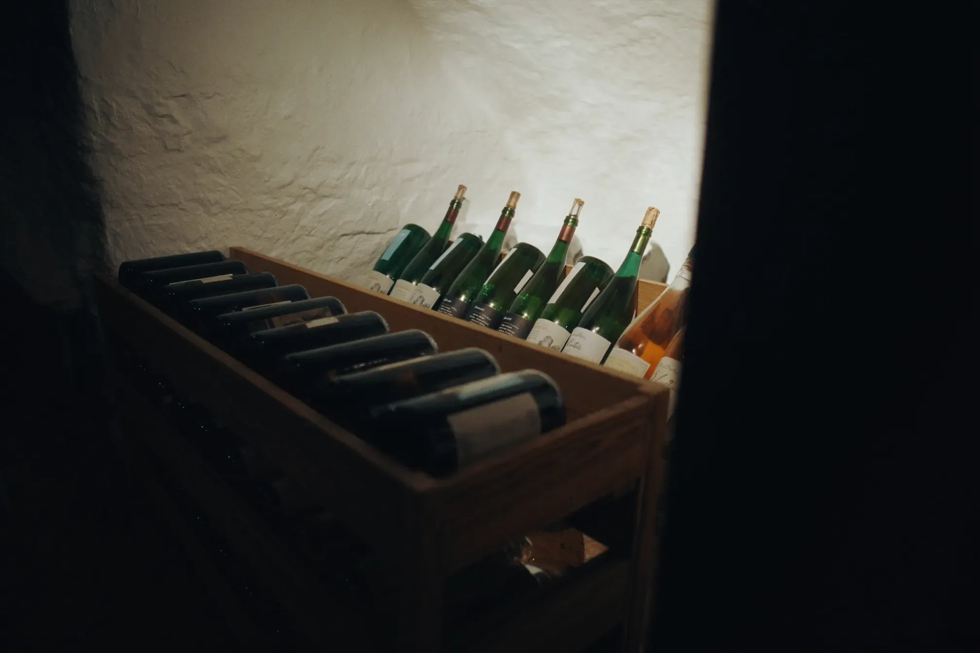 Dark wine bottles lying on their sides in a simple wooden rack, with a row of green bottles behind them against a whitewashed stone wall, lit by a single warm light in a dim cellar corner.