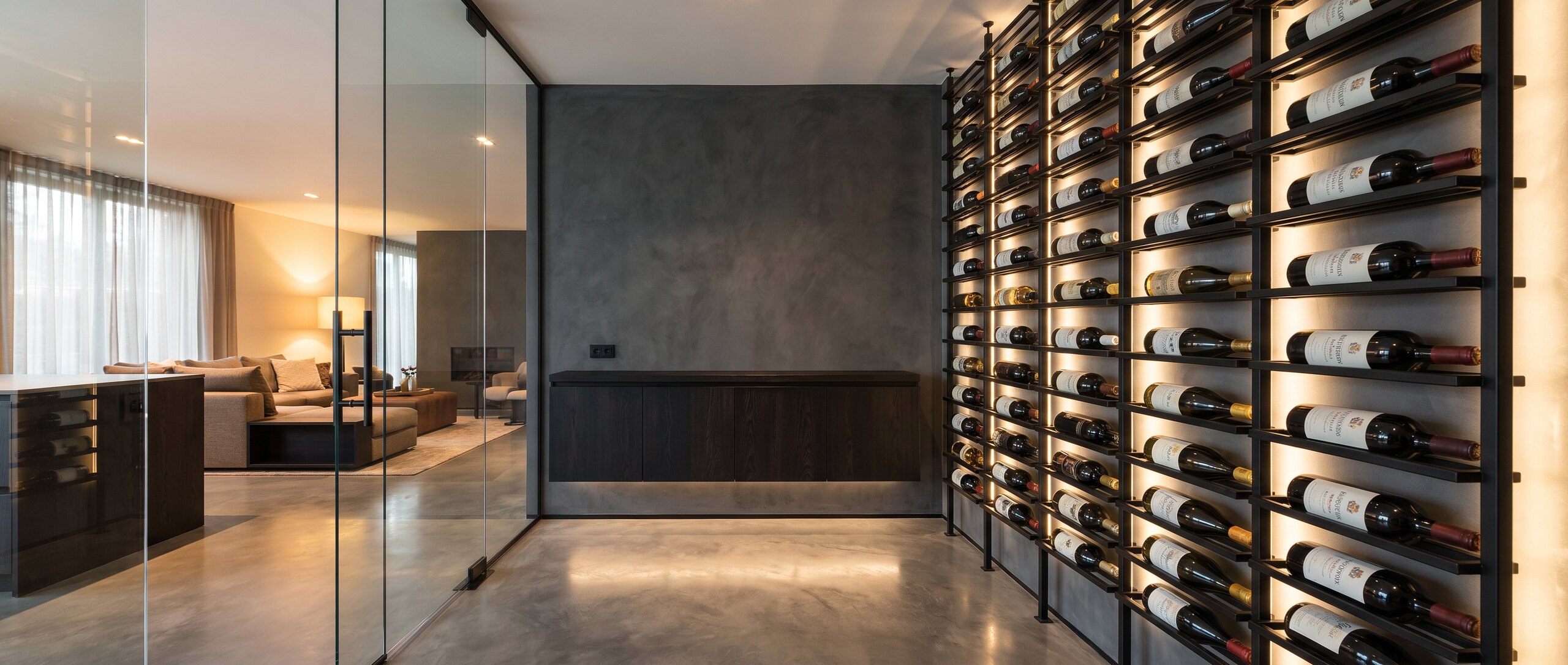 A sleek matte-black through-the-wall self-contained cooling unit recessed into a dark microcement wall, flanked by floor-to-ceiling matte-black metal wine racks, with a warm glass-walled tasting room alongside.