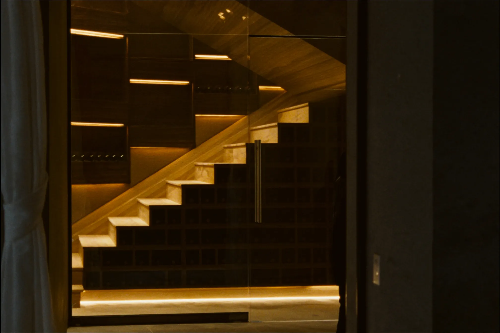 A wine cellar built under a staircase behind a frameless glass front, with wood racking following the slope of the stairs and warm LED strips under each tread.
